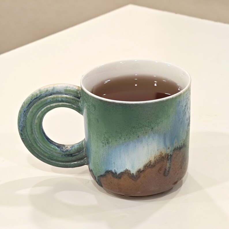 K&ouml;nnte beinhalten: Eine Keramik-Tasse mit einem einzigartigen Griff, gef&uuml;llt mit einer dunklen Fl&uuml;ssigkeit. Die Tasse hat einen Farbverlauf aus Gr&uuml;n, Blau und Braun. Das Innere ist wei&szlig;.