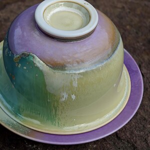 May include: A ceramic tea set with a purple, green, and yellow glaze. The set includes a teacup, a lid, and a saucer. The teacup is inverted, revealing a white rim and a light yellow interior.
