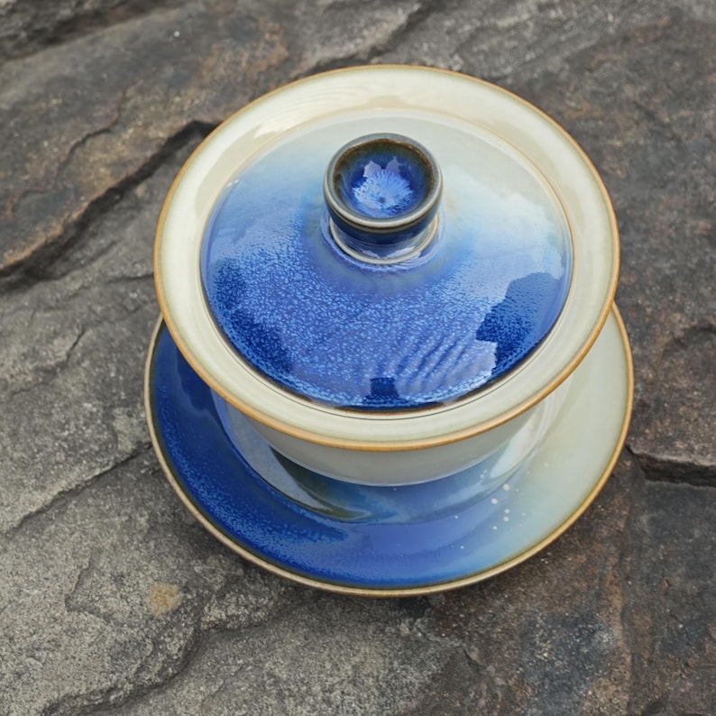 May include: A ceramic gaiwan tea set consisting of a bowl, a lid, and a saucer. The set features a gradient glaze transitioning from a deep, crystalline blue at the center to a creamy off-white at the edges, finished with a thin brown rim.