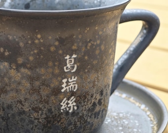 May include: A dark ceramic mug with a handle, on a matching saucer. The mug has a speckled, textured surface with white Japanese characters. The mug and saucer are dark grey with gold and white accents.