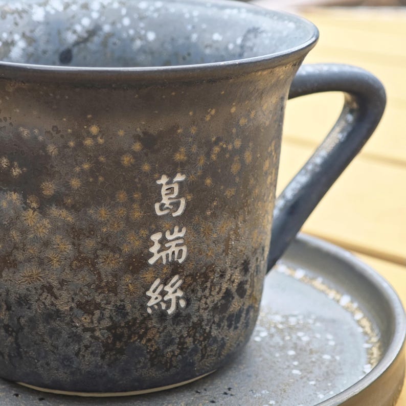 May include: A dark ceramic mug with a handle, resting on a matching saucer. The mug has a speckled, textured surface with white Japanese characters. The mug and saucer are dark gray with gold and white accents.