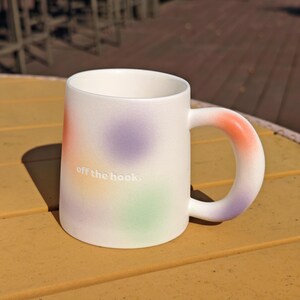 K&ouml;nnte beinhalten: Wei&szlig;e Keramiktasse mit einem farbenfrohen, gesprenkelten Design in Orange, Lila und Gr&uuml;n. Die Tasse hat einen abgerundeten Griff mit einem orangefarbenen Farbverlauf und der Schriftzug "off the hook." ist auf der Seite aufgedruckt.