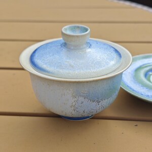 May include: A ceramic tea bowl with a lid, featuring a light blue and white glaze. The bowl has a rounded shape with a matching lid and a small handle. A matching saucer is partially visible in the background.