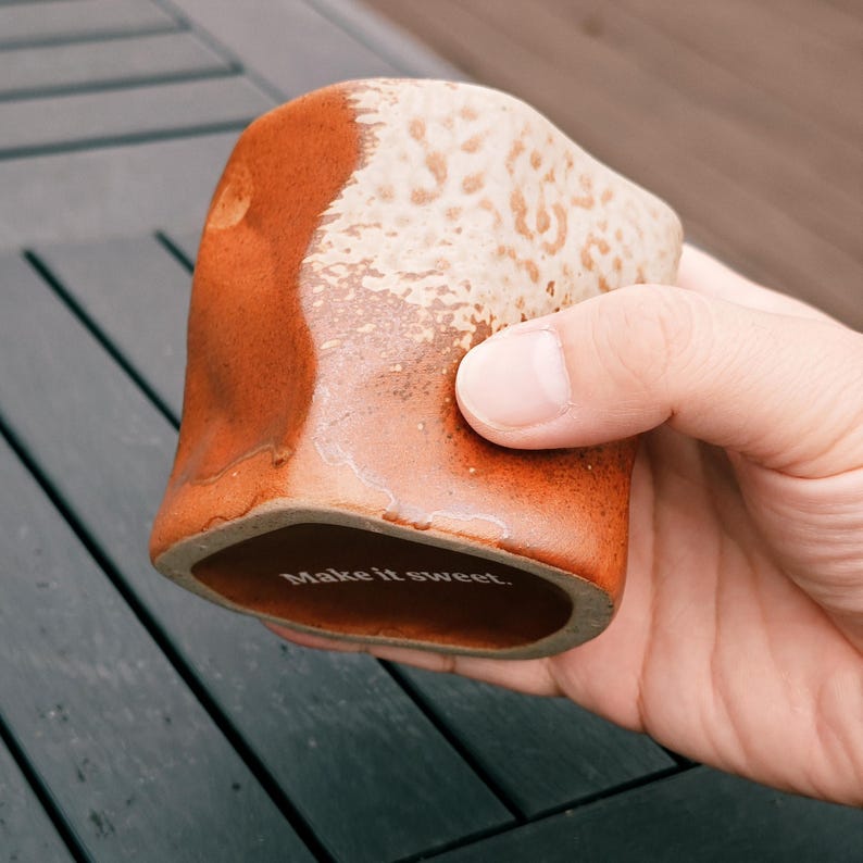 Peut inclure: Une tasse en c&eacute;ramique faite &agrave; la main avec un design textur&eacute; unique. La tasse pr&eacute;sente un m&eacute;lange de couleurs marron et cr&egrave;me, avec une base carr&eacute;e et un dessus incurv&eacute;. Le texte "Make it sweet" est imprim&eacute; au fond de la tasse.