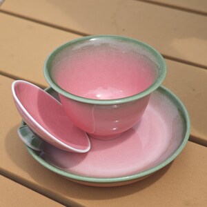 Puede incluir: Un juego de t&eacute; de cer&aacute;mica que consta de una taza en forma de cuenco, un platillo a juego y una tapa peque&ntilde;a. Las piezas presentan un interior y exterior de color rosa brillante, con un borde verde salvia que se difumina en el esmalte rosa.