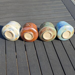 May include: Four ceramic mugs in a row, each with a unique color and inspirational text. The mugs are in shades of blue, brown, green, and blue. The text includes "Shine & Flow", "Breathe & Be", and "Dream & Create".
