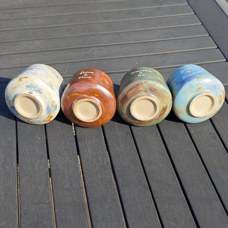 May include: Four ceramic mugs in a row, each with a unique color and inspirational text. The mugs are in shades of blue, brown, green, and blue. The text includes "Shine & Flow", "Breathe & Be", and "Dream & Create".
