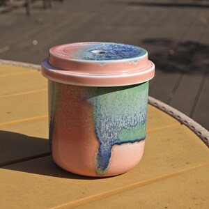 Pu&ograve; includere: Un barattolo in ceramica con coperchio. Il barattolo &egrave; cilindrico con base arrotondata. L'esterno presenta una miscela di colori, tra cui rosa, verde e blu. Il coperchio &egrave; rosa con un centro blu e verde. Il barattolo &egrave; appoggiato su una superficie gialla.