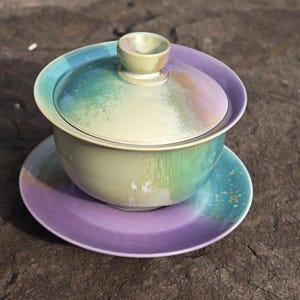 May include: A ceramic gaiwan tea set with a lid, bowl, and saucer. The set features a gradient of colors, including green, blue, and purple. The lid has a small handle, and the saucer is a solid purple color.