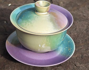 Puede incluir: Un juego de té gaiwan de cerámica con tapa, cuenco y platillo. El juego presenta un degradado de colores, incluyendo verde, azul y morado. La tapa tiene un pequeño asa y el platillo es de color morado liso.