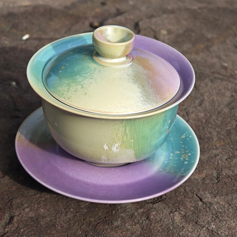 May include: A ceramic gaiwan tea set with a lid, bowl, and saucer. The set features a gradient of colors, including green, blue, and purple. The lid has a small handle, and the saucer is a solid purple color.