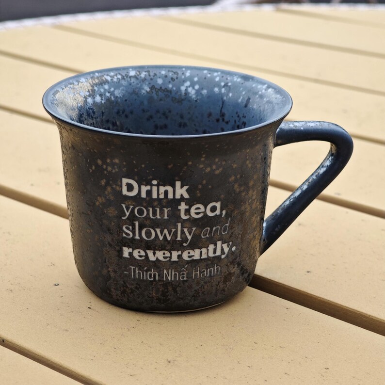 May include: A dark blue ceramic mug with a speckled pattern and a curved handle. The mug has the quote "Drink your tea, slowly and reverently" by Thich Nhat Hanh in white text. The mug is on a light brown surface.