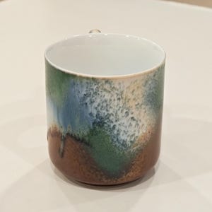 K&ouml;nnte beinhalten: Eine Keramiktasse mit einer einzigartigen, strukturierten Glasur. Die Tasse hat eine Farbpalette aus Braun, Gr&uuml;n, Blau und Wei&szlig; und einen kleinen Henkel. Der Innenraum ist wei&szlig; und steht im Kontrast zum k&uuml;nstlerischen Design des &Auml;u&szlig;eren. Ein dekoratives St&uuml;ck f&uuml;r die K&uuml;che.