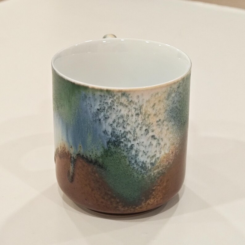 K&ouml;nnte beinhalten: Eine Keramiktasse mit einer einzigartigen, strukturierten Glasur. Die Tasse hat eine Farbpalette aus Braun, Gr&uuml;n, Blau und Wei&szlig; und einen kleinen Henkel. Der Innenraum ist wei&szlig; und steht im Kontrast zum k&uuml;nstlerischen Design des &Auml;u&szlig;eren. Ein dekoratives St&uuml;ck f&uuml;r die K&uuml;che.