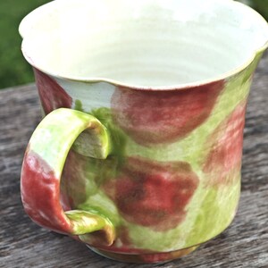 May include: A handmade ceramic mug with a unique, organic shape. The mug features a green and red mottled pattern on a cream-colored background. The handle is a vibrant green and red. The interior is a pale cream color.
