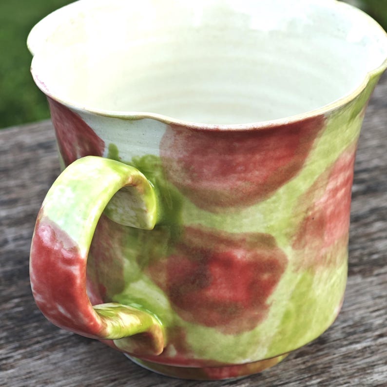 May include: A handmade ceramic mug with a unique, organic shape. The mug features a green and red mottled pattern on a cream-colored background. The handle is a vibrant green and red. The interior is a pale cream color.