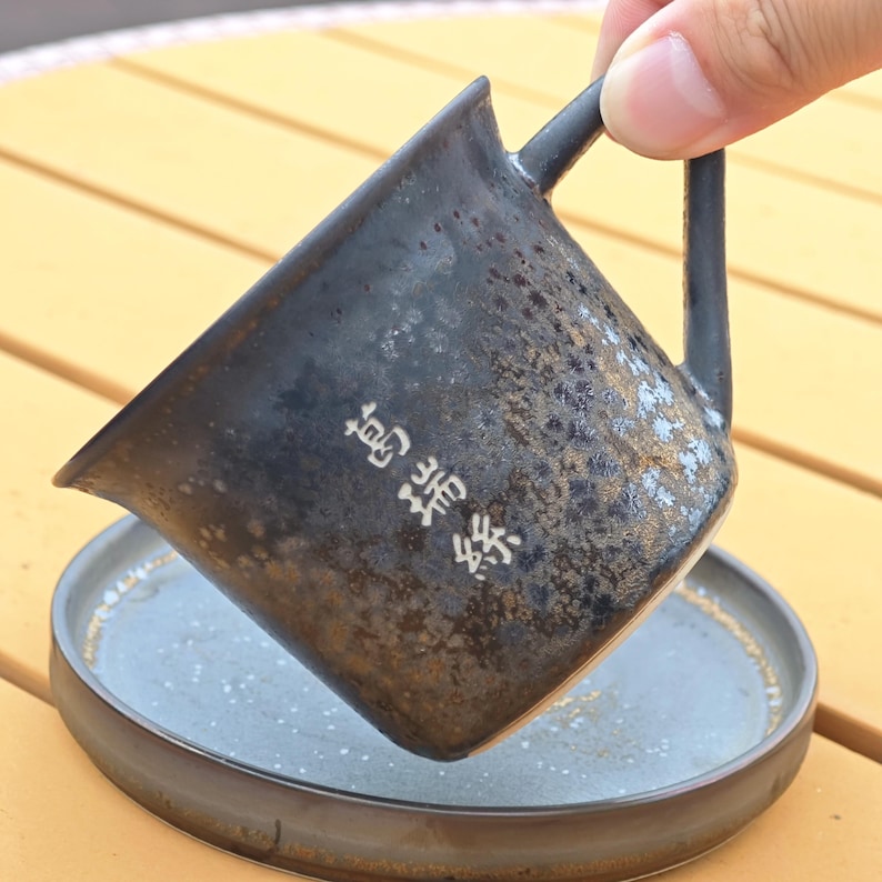 May include: A dark ceramic mug with a handle and a matching saucer. The mug has a textured, speckled surface with white Japanese characters. The saucer is a similar color and texture. The mug is being held over the saucer.