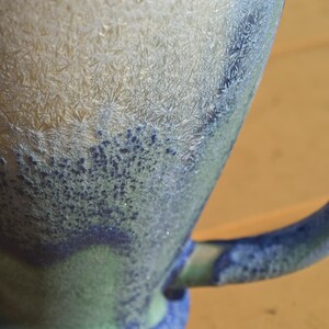 May include: Close-up of a ceramic mug with a textured, crystalline surface. The mug features a gradient of colors, transitioning from light beige to shades of blue and green. The handle is a deep blue.