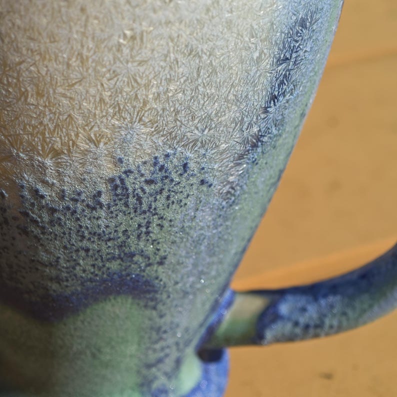 May include: Close-up of a ceramic mug with a textured, crystalline surface. The mug features a gradient of colors, transitioning from light beige to shades of blue and green. The handle is a deep blue.