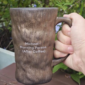 Puede incluir: Una taza de cer&aacute;mica marr&oacute;n con una superficie texturizada similar a la corteza. El frente presenta un texto impreso en blanco que dice: Michael - Morning Person (After Coffee). La taza tiene un asa resistente y una forma cil&iacute;ndrica.