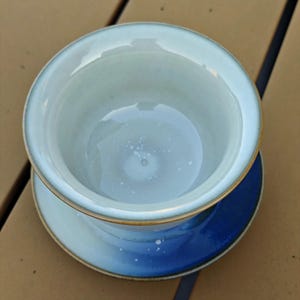 May include: A ceramic bowl with a light, pale blue interior and a smooth, glossy finish. The exterior features a gradient transition from a light blue at the top to a deep, saturated blue at the base, with a thin, dark brown rim around the edge.
