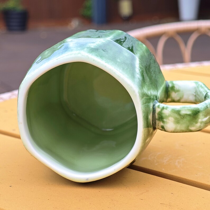 May include: A green ceramic mug with a geometric design. The mug has a white rim and handle. The interior of the mug is a lighter shade of green. The mug is sitting on a yellow surface.