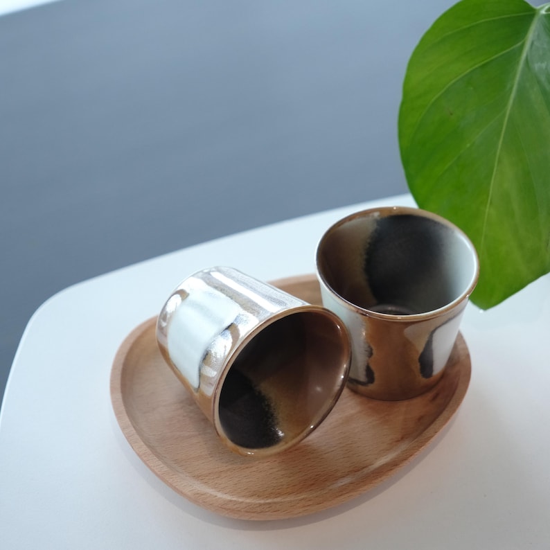 以下が含まれることがあります： 木製のトレイに、茶色、白、黒の釉薬を施した2つのセラミックカップが置かれています。カップは円筒形で、滑らかで光沢のある仕上がりです。トレイは楕円形で、明るい色の木材で作られています。