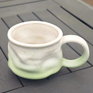 Op de afbeelding: Een handgemaakte keramische mok met een witte en groene ombre glazuur. De mok heeft een afgeronde vorm met een grote handgreep en een gespikkeld interieur. De buitenkant heeft een getextureerd, organisch gevoel.