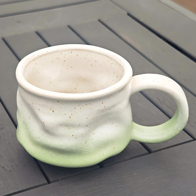 Op de afbeelding: Een handgemaakte keramische mok met een witte en groene ombre glazuur. De mok heeft een afgeronde vorm met een grote handgreep en een gespikkeld interieur. De buitenkant heeft een getextureerd, organisch gevoel.