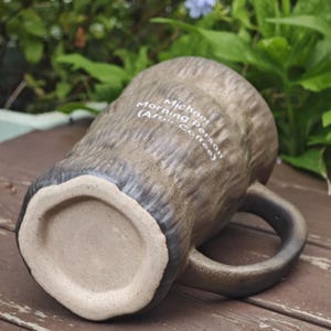 Puede incluir: Una taza de cer&aacute;mica marr&oacute;n con un exterior texturizado similar a la corteza y una base lisa de color claro. La taza tiene un asa oscura y texto impreso en blanco que dice: Michael Morning Person (After Coffee). Est&aacute; colocada de lado.