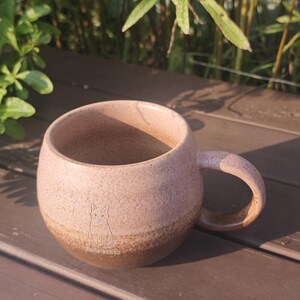 Może przedstawiać: Okrągły ceramiczny kubek z nakrapianą r&oacute;żową g&oacute;rną częścią i jednolitym brązowym dnem. Na przodzie kubka znajduje się mały, wygrawerowany rysunek liniowy kota, umieszczony nad napisem "Hello Bunny" wykonanym prostą, cienką czcionką bezszeryfową.