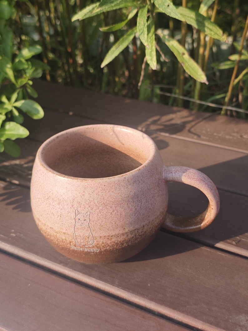 Może przedstawiać: Okrągły ceramiczny kubek z nakrapianą r&oacute;żową g&oacute;rną częścią i jednolitym brązowym dnem. Na przodzie kubka znajduje się mały, wygrawerowany rysunek liniowy kota, umieszczony nad napisem "Hello Bunny" wykonanym prostą, cienką czcionką bezszeryfową.