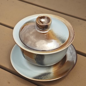 May include: A ceramic gaiwan tea set consisting of a bowl, a lid with a central knob, and a saucer. The pieces feature a light blue glaze with blended brown and charcoal grey accents, creating a smooth, glossy finish across the entire surface.