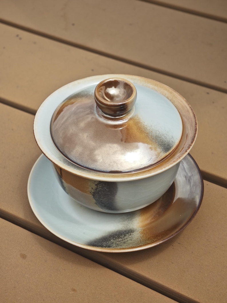 May include: A ceramic gaiwan tea set consisting of a bowl, a lid with a central knob, and a saucer. The pieces feature a light blue glaze with blended brown and charcoal grey accents, creating a smooth, glossy finish across the entire surface.