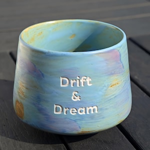 May include: A ceramic tumbler with a blue and gold glaze, featuring the words "Drift & Dream" etched into the side. The cup has a wide opening and a rounded base. The tumbler is sitting on a dark wooden surface.