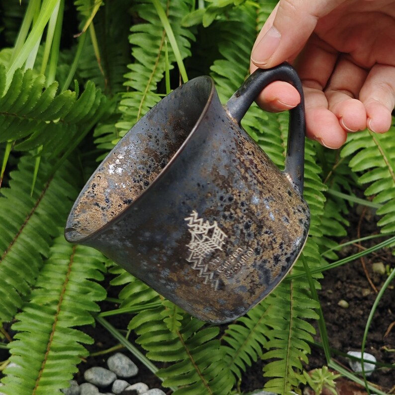 May include: A dark blue ceramic mug with a speckled texture and a black handle. The interior has a golden hue. The mug features an Asian-inspired design and text. The mug is held against a backdrop of green ferns.