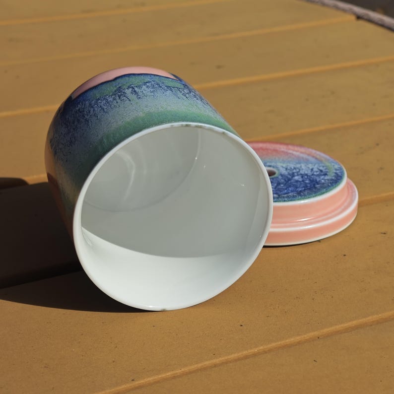 Pu&ograve; includere: Un barattolo di ceramica con coperchio. L'interno del barattolo &egrave; bianco e ha un esterno colorato con sfumature blu, verdi e rosa. Il coperchio &egrave; rosa con un disegno blu e verde. Il barattolo &egrave; sdraiato su un lato.
