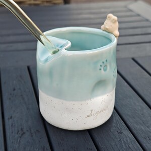 Puede incluir: Una taza de cerámica azul claro con una base blanca moteada. La taza presenta diseños de huellas de patas y una pequeña figura de perro en el borde. Una cuchara está insertada en la taza. La palabra "Sigma" está escrita en la base.