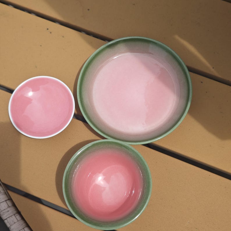 Puede incluir: Un juego de tres platos de cer&aacute;mica que consta de un platillo rosa peque&ntilde;o con borde blanco, un cuenco mediano con interior rosa y borde exterior verde, y un cuenco grande con interior rosa y borde exterior verde.