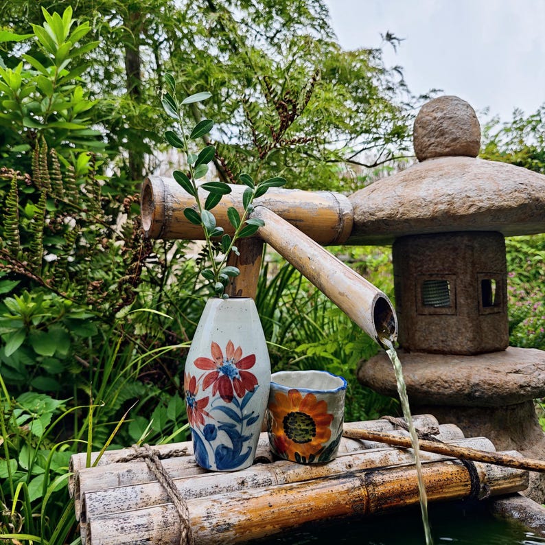 May include: A serene outdoor scene features a bamboo water fountain with water flowing into a small pond. A ceramic vase with red and blue floral designs holds a leafy branch. A small cup with a sunflower design sits nearby.