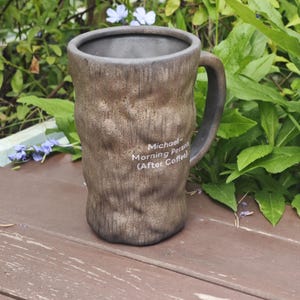 Puede incluir: Una taza de cer&aacute;mica cil&iacute;ndrica y alta con un acabado exterior marr&oacute;n texturizado similar a la corteza y un interior oscuro. La taza cuenta con un asa curva y texto impreso en blanco que dice: Michael Morning Person (After Coffee).