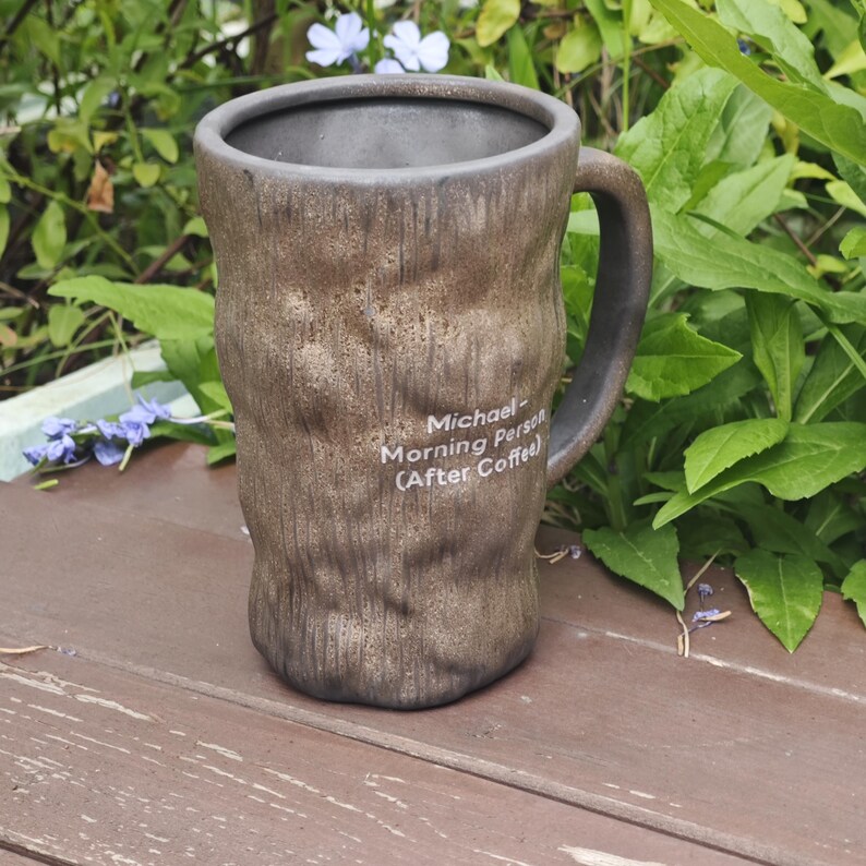 Puede incluir: Una taza de cer&aacute;mica cil&iacute;ndrica y alta con un acabado exterior marr&oacute;n texturizado similar a la corteza y un interior oscuro. La taza cuenta con un asa curva y texto impreso en blanco que dice: Michael Morning Person (After Coffee).