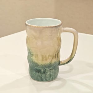May include: A tall, textured ceramic mug with a handle. The mug features a gradient of colors, transitioning from a light beige at the top to a teal green at the bottom. The interior is a light blue color.