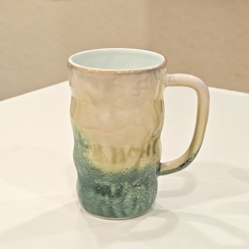 May include: A tall, textured ceramic mug with a handle. The mug features a gradient of colors, transitioning from a light beige at the top to a teal green at the bottom. The interior is a light blue color.