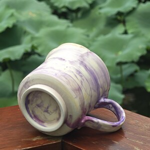 May include: A ceramic mug with a marbled design in shades of purple and cream. The mug has a rounded shape with a handle and is set against a blurred green background. The mug is resting on a wooden surface.