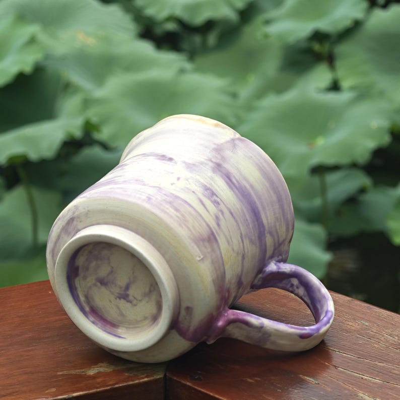 May include: A ceramic mug with a marbled design in shades of purple and cream. The mug has a rounded shape with a handle and is set against a blurred green background. The mug is resting on a wooden surface.