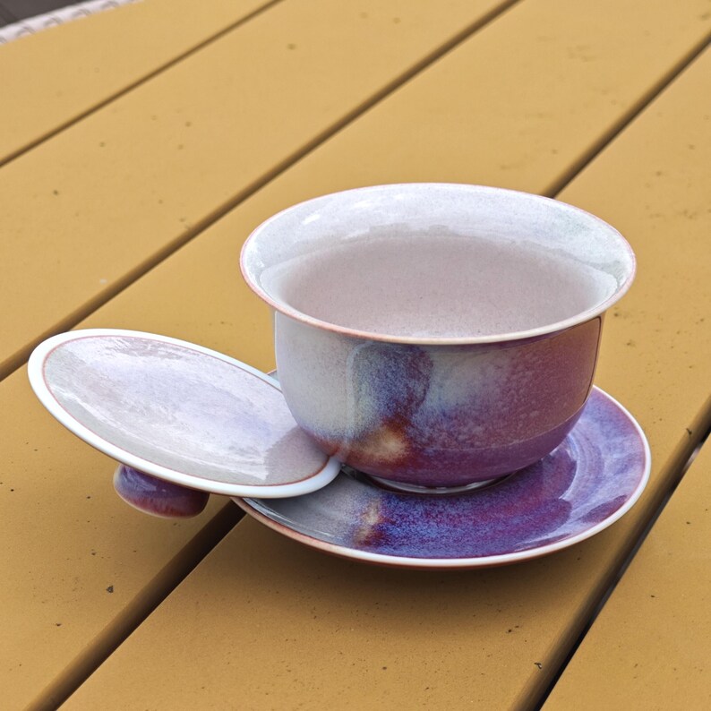 May include: A ceramic tea set with a purple and white glaze. The set includes a teacup, saucer, and lid. The teacup and saucer are round, and the lid is oval. The set is designed for tea drinking.