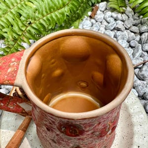 Op de afbeelding: Een handgemaakte keramische mok met een getextureerde, roodbruine buitenkant en een lichtbruine binnenkant. De handgreep en de buitenkant hebben een gespikkeld ontwerp. De mok staat op een wit gespikkelde plaat, met een kaneelstokje en varenbladeren op de achtergrond.
