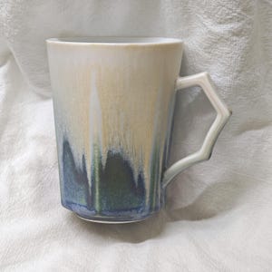 May include: A tall ceramic mug with a unique handle design. The mug features a gradient of colors, transitioning from a light beige at the top to a deep blue at the base. The handle has an angular, geometric shape.