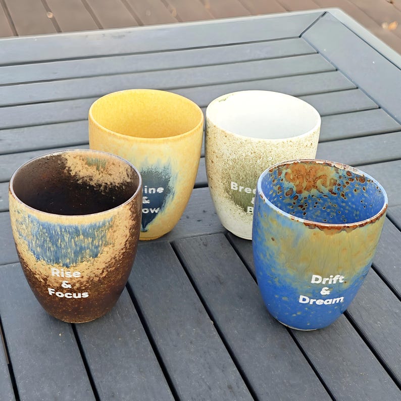 Puede incluir: Cuatro tazas de cer&aacute;mica con un acabado moteado y esmaltado en tonos marr&oacute;n, amarillo, azul y blanco. Cada taza tiene un dise&ntilde;o &uacute;nico y el texto: "Rise & Focus", "Shine & Glow", "Breathe & Be" y "Drift & Dream".
